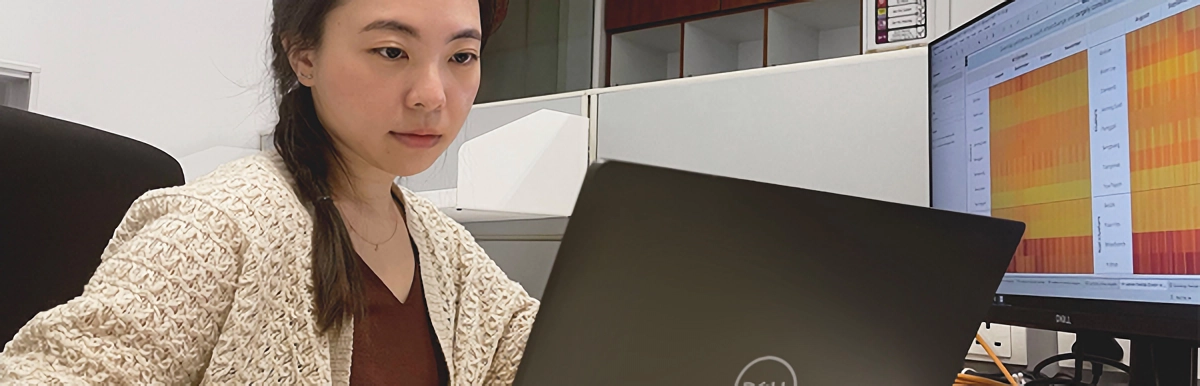 Young woman working at a Dell laptop with a monitor displaying a colorful data heatmap behind her.
